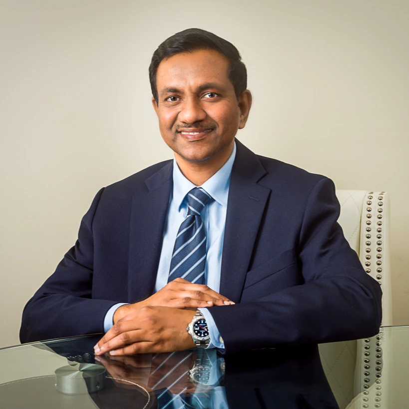 Man in dark suit and blue tie sitting at desk, smiling at camera