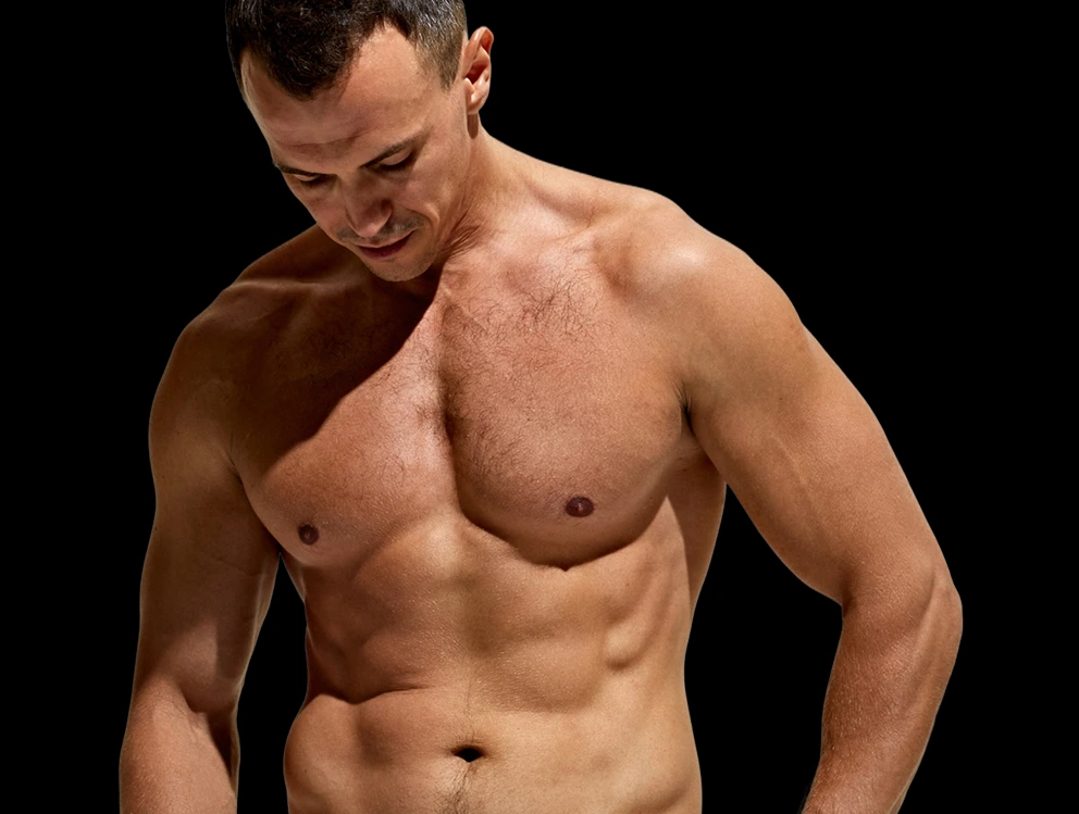 Shirtless man with defined chest and abdominal muscles looking downward against black background